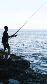 A man fishes on a rocky seashore at sunrise, enjoying a peaceful morning by the ocean.